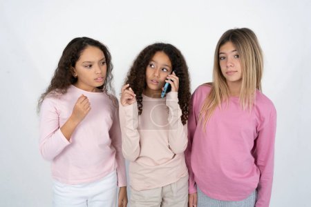 Three young beautiful multiracial kid girls speaks on mobile phone spends free time indoors calls to friend.