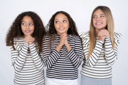 Téléchargez les photos : Heureux Trois jeunes belles filles multiraciales enfants anticipe quelque chose d'impressionnant se produire, regarde heureusement de côté, garde les mains ensemble près du visage, a une expression heureuse. - en image libre de droit