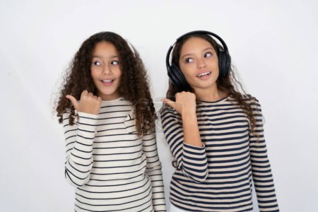 Téléchargez les photos : Deux jeunes belles filles multiraciales enfants écoute la piste audio via casque sans fil points pouce loin annonce l'espace de copie - en image libre de droit