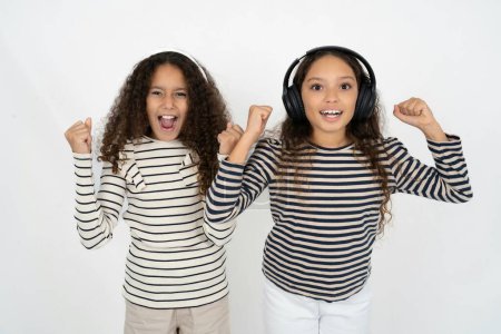Photo for Emotional two young beautiful multiracial kid girls exclaims loudly feels like winner raises clenched fists keeps mouth opened wears stereo headphones on ears makes yes gesture, listens favorite music - Royalty Free Image