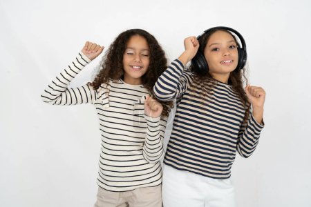 Photo for Carefree two young beautiful multiracial kid girls with toothy smile raises arms dances carefree moves with rhythm of music listens music from playlist via headphones - Royalty Free Image