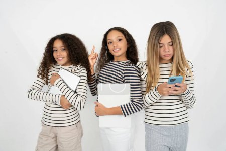 Téléchargez les photos : Trois jeunes belles filles multiraciales enfant tenant ordinateur portable montrant copie vide espace publicitaire - en image libre de droit