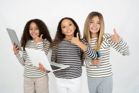 Photo for Beautiful attractive Three young beautiful multiracial kid girls hold netbook hand finger show thumb up - Royalty Free Image
