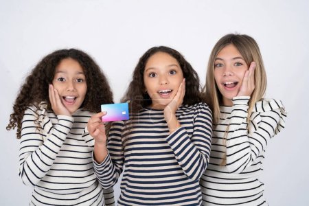 Foto de Sorprendido positivo Tres jóvenes hermosas niñas multirraciales brazo cara sostener banco tarjeta de plástico funciones impresas - Imagen libre de derechos