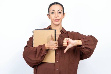 Photo for Beautiful woman arab student carries notebooks with paper showing thumbs up and thumbs down, difficult choose concept - Royalty Free Image
