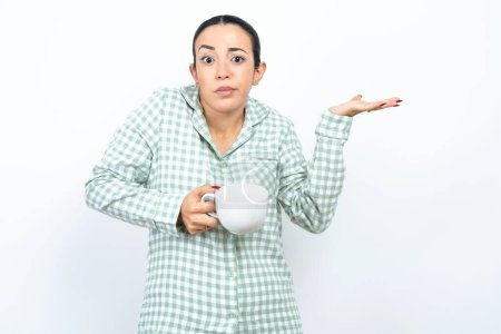Téléchargez les photos : Belle jeune femme portant un pyjama à carreaux verts et tenant une tasse pointant de côté avec les deux mains montrant quelque chose d'étrange et disant : Je ne sais pas ce que c'est. Concept de publicité. - en image libre de droit