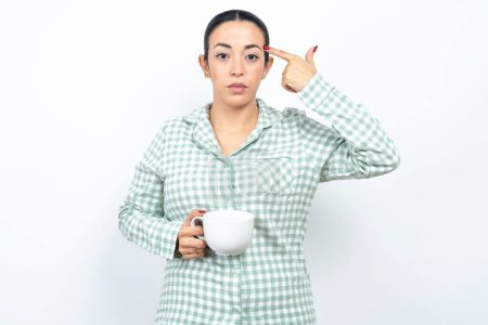 Photo for Unhappy Beautiful young woman wearing green plaid pyjama and holding a cup makes suicide gesture and imitates gun with hand, curves lips keeps two fingers on temple, shoots, being tired of everything, - Royalty Free Image