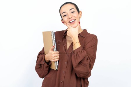Foto de Hermosa estudiante árabe mujer lleva cuadernos con papel mirando con confianza a la cámara sonriendo con los brazos cruzados y la mano levantada en la barbilla. Pensando positivamente. - Imagen libre de derechos