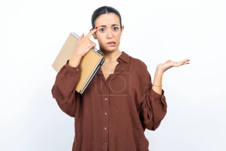 Photo for Beautiful woman arab student carries notebooks with paper confused and annoyed with open palm showing copy space and pointing finger to forehead. Think about it. - Royalty Free Image