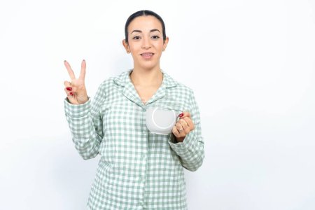 Photo for Beautiful young woman wearing green plaid pyjama and holding a cup showing and pointing up with fingers number two while smiling confident and happy. - Royalty Free Image