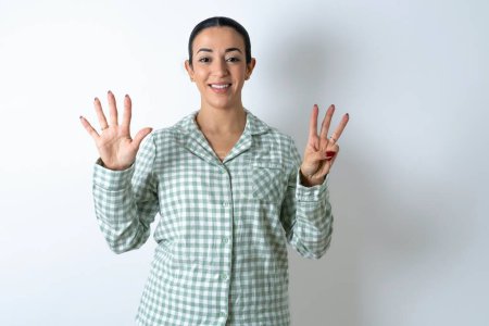 Foto de Hermosa joven con pijama a cuadros verde sobre fondo blanco mostrando y apuntando hacia arriba con los dedos número ocho mientras sonríe confiado y feliz. - Imagen libre de derechos