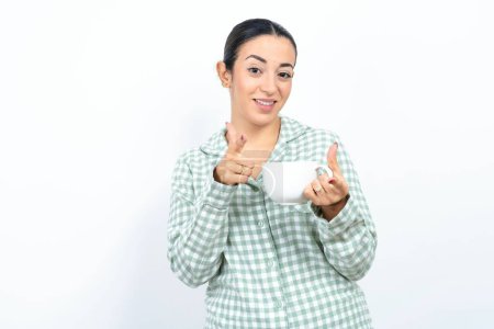Foto de Hermosa mujer joven con pijama a cuadros verde y sosteniendo una taza señalando los dedos a la cámara con la cara feliz y divertida. Buena energía y vibraciones. - Imagen libre de derechos