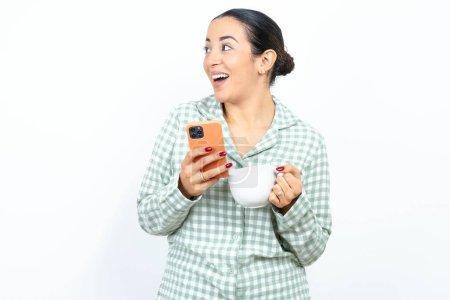 Téléchargez les photos : Belle jeune femme portant un pyjama à carreaux vert et tenant une tasse tenant un smartphone et regardant de côté l'espace de copie vierge. - en image libre de droit