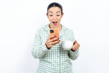 Foto de Hermosa mujer joven con pijama a cuadros verde y sosteniendo una taza usando el teléfono móvil charlando tiempo libre . - Imagen libre de derechos