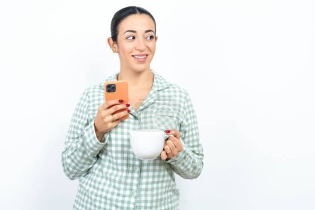 Photo for Beautiful young woman wearing green plaid pyjama and holding a cup hold telephone hands read good youth news look empty space advert - Royalty Free Image