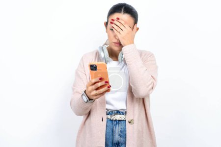 Téléchargez les photos : Belle femme arabe posant sur fond blanc regardant téléphone intelligent se sentant triste tenant la main sur le visage. - en image libre de droit