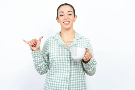 Téléchargez les photos : Belle jeune femme portant pyjama à carreaux vert et tenant une tasse montrant le numéro six Liu avec geste des doigts en langue chinoise des signes - en image libre de droit
