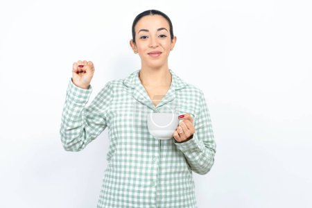Foto de Hermosa joven con pijama a cuadros verde y sosteniendo una taza apuntando hacia arriba con los dedos número diez en chino Shi lenguaje de signos - Imagen libre de derechos