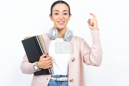Téléchargez les photos : Belle étudiante arabe porte des cahiers sur fond blanc pointant vers le haut avec les doigts numéro neuf en langue des signes chinoise Jiu - en image libre de droit