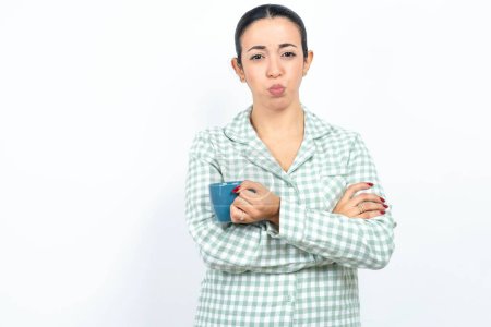 Gloomy dissatisfied Beautiful young woman wearing green plaid pyjama and holding a cup looks with miserable expression at camera from under forehead, makes unhappy grimace