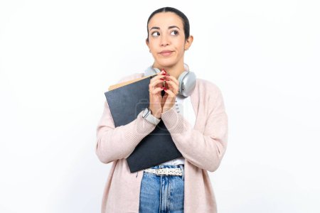 Téléchargez les photos : Belle étudiante arabe porte des cahiers sur fond blanc doigts raides et semble mystérieux de côté a grand plan mal à l'esprit - en image libre de droit