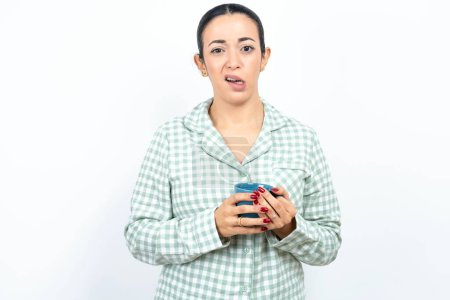Photo for Portrait of dissatisfied Beautiful young woman wearing green plaid pyjama and holding a cup smirks face, purses lips and looks with annoyance at camera, discontent hearing something unpleasant - Royalty Free Image