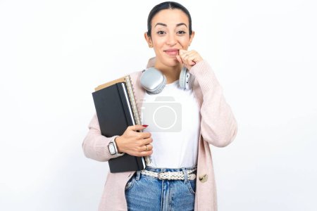 Photo for Pleased beautiful arab woman student carries notebooks over white background with closed eyes keeps hands near cheeks and smiles tenderly imagines something very pleasant - Royalty Free Image