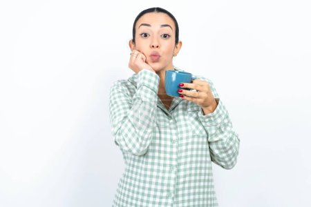 Photo for Beautiful young woman wearing green plaid pyjama and holding a cup with surprised expression keeps hands under chin keeps lips folded makes funny grimace - Royalty Free Image