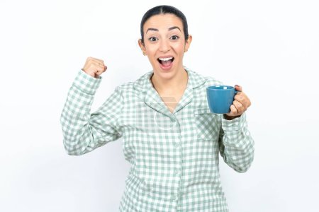 Photo for Shocked ecstatic Beautiful young woman wearing green plaid pyjama and holding a cup win luck lottery raise hands up shout yea - Royalty Free Image