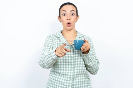 Photo for Shocked Beautiful young woman wearing green plaid pyjama and holding a cup points at you with stunned expression - Royalty Free Image