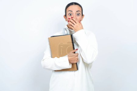 Photo for Stunned beautiful arab woman student carries notebooks over white background covers both hands on mouth, afraids of something astonishing - Royalty Free Image