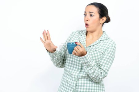 Téléchargez les photos : Mécontent Belle jeune femme portant un pyjama à carreaux vert et tenant une tasse garde les mains vers l'espace vide et demande de ne pas se rapprocher voit quelque chose de désagréable - en image libre de droit