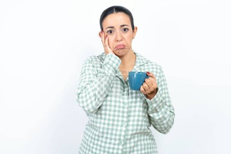 Photo for Sad lonely Beautiful young woman wearing green plaid pyjama and holding a cup touches cheek with hand bites lower lip and gazes with displeasure. Bad emotions - Royalty Free Image