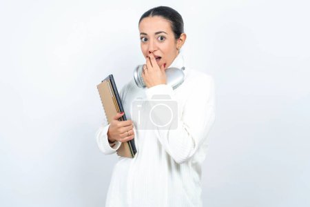 Téléchargez les photos : Belle étudiante arabe porte des cahiers sur fond blanc couvre la bouche et regarde avec émerveillement à la caméra, ne peut pas croire rumeurs inattendues. - en image libre de droit