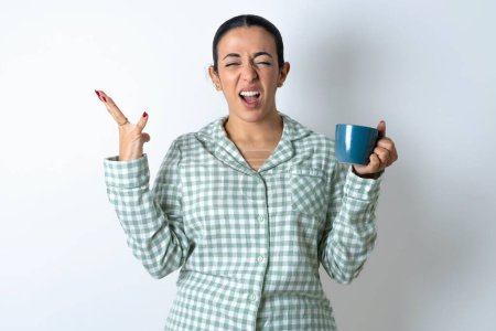 Téléchargez les photos : Folle indignée Belle jeune femme portant un pyjama à carreaux vert et tenant une tasse crie bruyamment et se met à hurler furieusement. Négatif émotions humaines sentiments concept - en image libre de droit