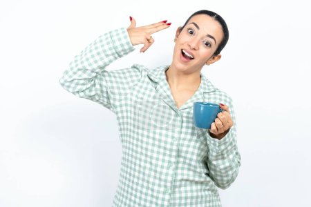 Photo for Beautiful young woman wearing green plaid pyjama and holding a cup foolishness around shoots in temple with fingers makes suicide gesture. Funny model makes finger gun pistol - Royalty Free Image