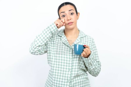 Photo for Disappointed dejected Beautiful young woman wearing green plaid pyjama and holding a cup wipes tears stands stressed with gloomy expression. Negative emotion - Royalty Free Image