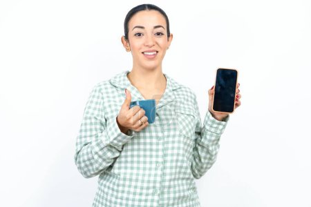 Foto de Hermosa mujer joven con pijama a cuadros verde y sosteniendo una taza Mostrar teléfono inteligente de pantalla en blanco, pulgar hacia arriba recomiendan nueva aplicación - Imagen libre de derechos