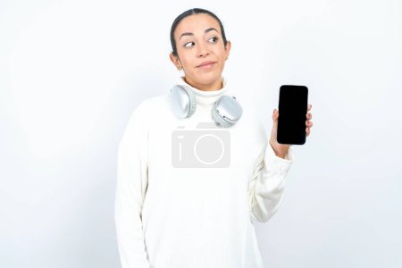 Téléchargez les photos : Belle femme arabe posant sur fond blanc tient un nouveau téléphone mobile et semble mystérieux montre l'affichage vierge de cellulaire moderne - en image libre de droit