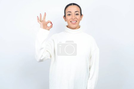 Foto de Hermosa mujer árabe posando sobre fondo blanco mano brazo okey símbolo toothy aprobar asesorar novedad noticias - Imagen libre de derechos