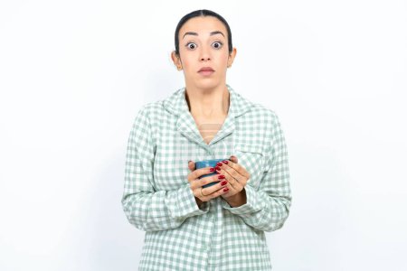 Photo for Stunned Beautiful young woman wearing green plaid pyjama and holding a cup stares reacts on shocking news. Astonished MODEL holds breath - Royalty Free Image