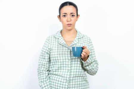 Téléchargez les photos : Belle jeune femme portant pyjama à carreaux vert et tenant une tasse pointant vers le bas avec les doigts montrant la publicité, visage surpris et bouche ouverte - en image libre de droit