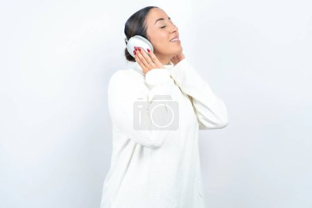 Téléchargez les photos : Belle femme arabe posant sur fond blanc avec écouteurs sur la tête, écoute de la musique, profiter de la chanson préférée avec les yeux fermés, tenant la main sur casque. - en image libre de droit