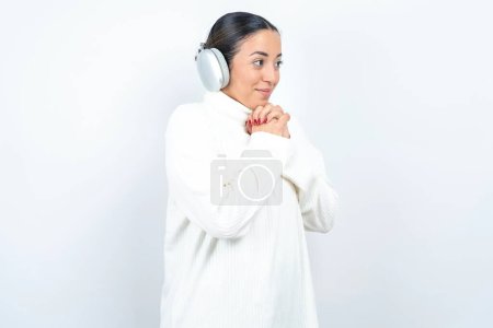 Foto de Hermosa mujer árabe posando sobre fondo blanco lleva auriculares estéreo escuchando música concentrada y mirando a un lado con interés. - Imagen libre de derechos