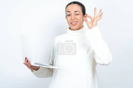Téléchargez les photos : Attrayant joyeux qualifié belle femme arabe posant sur fond blanc en utilisant un ordinateur portable montrant ok-signe winkin - en image libre de droit