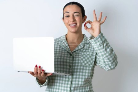 Foto de Positivo Hermosa mujer joven con pijama a cuadros verde sobre fondo blanco mantenga los dedos inalámbricos netbook mano muestran símbolo okey - Imagen libre de derechos