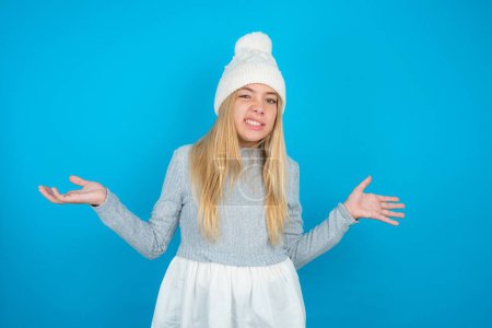 Photo for Clueless Beautiful kid girl wearing white knitted hat and blue sweater shrugs shoulders with hesitation, faces doubtful situation, spreads palms, Hard decision - Royalty Free Image