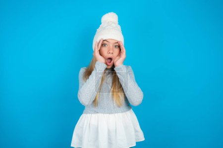 Photo for Beautiful kid girl wearing white knitted hat and blue sweater with scared expression, keeps hands on head, jaw dropped, has terrific expression. Omg concept - Royalty Free Image