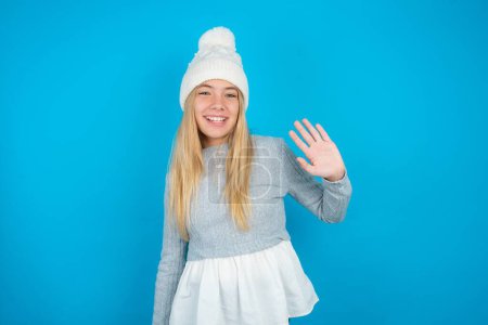 Foto de Hermosa niña con sombrero de punto blanco y suéter azul Renuncia a decir hola feliz y sonriente, gesto de bienvenida amistoso. - Imagen libre de derechos