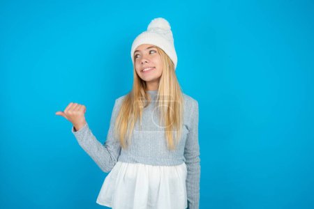 Foto de Hermosa niña encantadora con sombrero de punto blanco y suéter azul mirando espacio de copia que tiene anuncios - Imagen libre de derechos